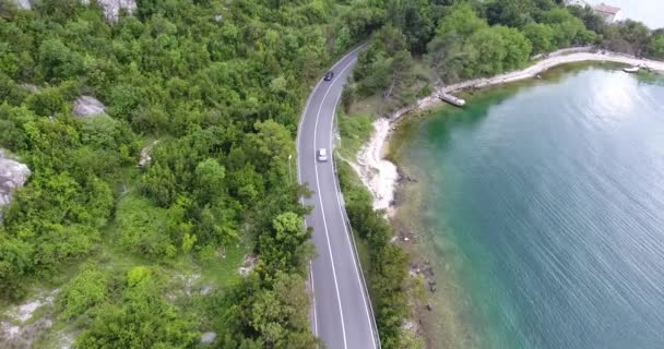 四轴飞行器正飞过一条狭窄的柏油路 沿着海岸行驶 我们可以看到绿松石的大海和海岸覆盖着绿色的树木在道路的另一边 卡车和旅游巴士沿路行驶 — 图库视频影像