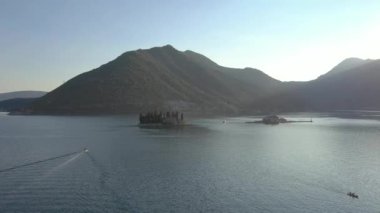 Adriyatik kıyıları üzerinde uçan, gök mavisi deniz ve üzerinde taş evler ve kilise olan küçük bir ada tatil köyü. Arka planda dağlar görünür.