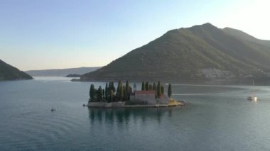 Adriyatik kıyıları üzerinde uçan, gök mavisi deniz ve üzerinde kozalaklı ağaçlar, taş evler ve kilise bulunan küçük bir ada tatil köyü. Arka planda dağlar görünür.