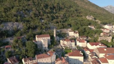 Adriyatik kıyısındaki küçük Karadağ kenti Perast üzerinde uçuş. Beyaz taş evlerin ve ağaçların çatıları olan antik bir kilise görebiliyoruz. Gün batımı. Arkaplanda dağlar