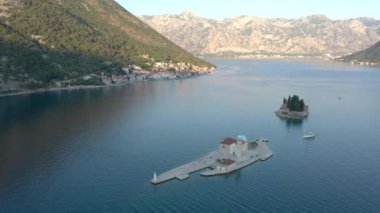 Karadağ 'ın Adriyatik denizinde küçük bir adanın üzerinde uçuyor. Gök mavisi deniz ve üzerinde kozalaklı ağaçlar, beyaz taş evler ve resifteki Bakire Kilisesi olan küçük bir ada. Arka planda dağlar görünür.