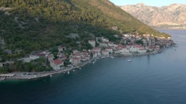 Adriyatik kıyısındaki küçük Karadağ kenti Perast üzerinde uçuş. Beyaz taş evlerin ve ağaçların çatıları olan antik bir kilise görebiliyoruz. Gün batımı. Arkaplanda dağlar