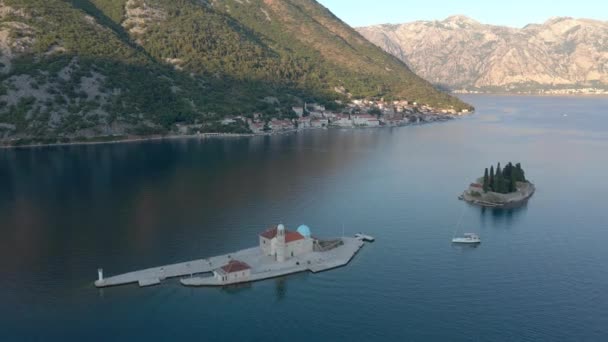 飞越了位于黑山亚得里亚海的一个小岛 蓝色的大海和一个小岛 岛上有针叶树 白色的石头房子和岛上的圣母教堂 背景中可以看到山脉 — 图库视频影像
