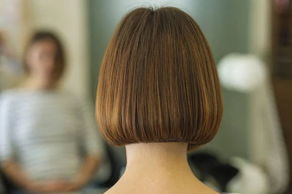 Young woman had her hair cut and styled in a beauty salon. She is sitting facing the mirror, and from behind we can see a straight line of her brown hair reaching up to the neck