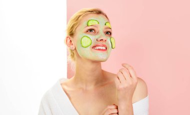 Yüzünde kil kozmetik maske ve taze salatalık dilimleri olan gülümseyen genç bir kadın. Spa salonu, güzellik tedavisi, kozmetik prosedürler. Kozmetoloji ve cilt bakımı. Salatalık maskesi. Doğal malzemeler.
