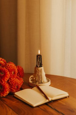 Ahşap masada yanan mum, defter, kalem ve portakallı yıldız çiçekleriyle dolu sıcak bir sonbahar hayatı. Sıcak ev dekoru, sonbahar havası, hygge atmosferi, rahatlama ve farkındalık konsepti..