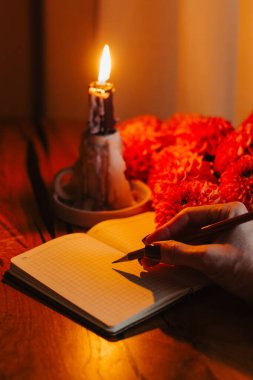 Sıcak bir akşam sahnesi, yanan bir mum, kırmızı çiçekler ve açık bir defterin üzerinde kalem tutan bir el, samimi ve ilham verici bir yazı atmosferi yaratıyor..
