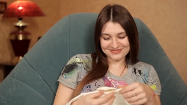 Счастливая беременная девушка сидит в кресле у камина и смотрит на одежду для новорожденных — стоковое видео