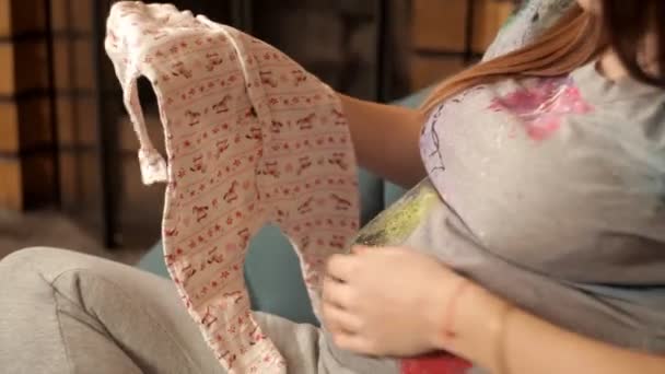 Menina grávida feliz senta-se em uma poltrona perto da lareira e olha para a roupa para recém-nascidos — Vídeo de Stock