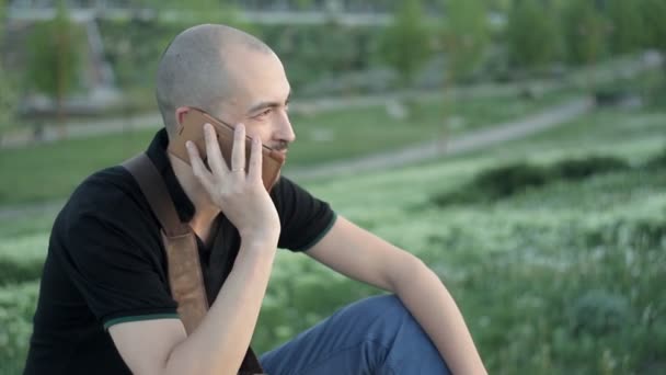 Joven sonriente sentado en el parque con una bolsa en el fondo de la vegetación y hablando por teléfono — Vídeos de Stock