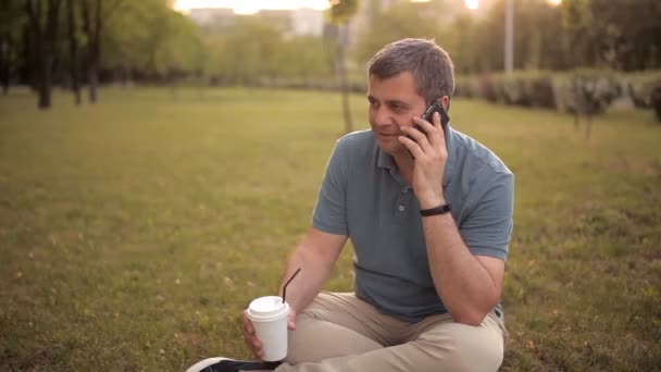 Un hombre sentado en el césped en un parque verde en un día de verano al atardecer bebiendo una bebida y hablando por teléfono — Vídeo de stock