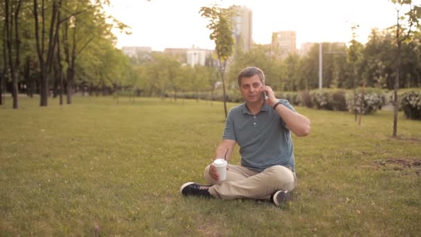 Мужчина, сидящий на лужайке в зеленом парке в летний день на закате, пьющий напиток и разговаривающий по телефону — стоковое видео