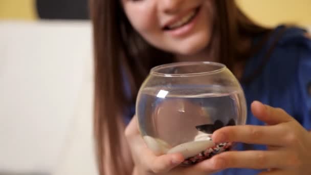 Молодая улыбающаяся девушка в синем костюме сидит на диване и стучится в маленький аквариум с рыбой и наблюдает, как рыба плавает — стоковое видео