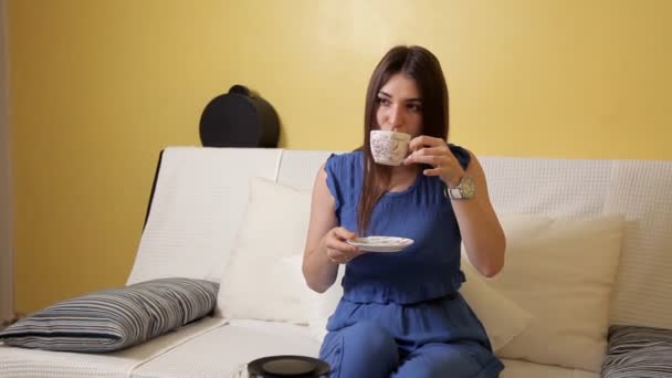 Ένα νεαρό κορίτσι σε ένα μπλε κοστούμι κάθεται στον καναπέ και πίνει τσάι σκέφτεται κάτι. Πίνοντας τσάι — Αρχείο Βίντεο
