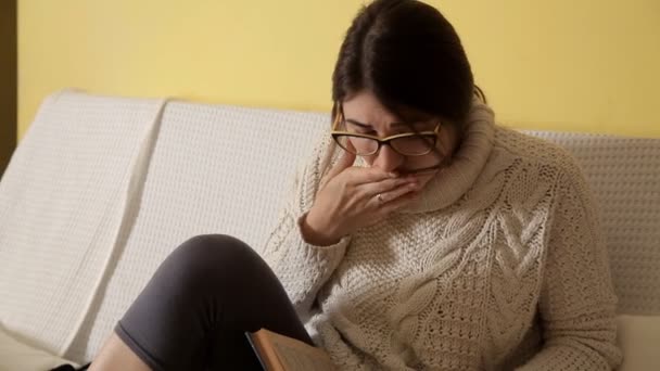 Ένα νεαρό κορίτσι σε ένα λευκό πουλόβερ για ένα χειμώνα στο σπίτι το βράδυ με γυαλιά, διαβάζοντας ένα βιβλίο. Βιβλίο. Το βράδυ. Κορίτσι. ανάγνωση. Συναισθήματα — Αρχείο Βίντεο