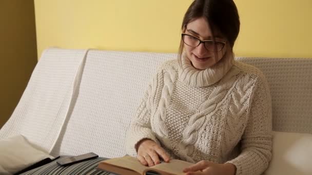 Une jeune fille en pull blanc un soir d'hiver à la maison avec des lunettes lit un livre, appelle par téléphone, une fille parle au téléphone. Livre. Bonsoir. Fille. lecture. Émotion.Se reposer — Video