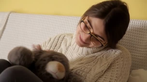 Een jong meisje in een witte trui en dragen bril wordt gespeeld met een zacht stuk speelgoed. Jeugd. Speelgoed — Stockvideo