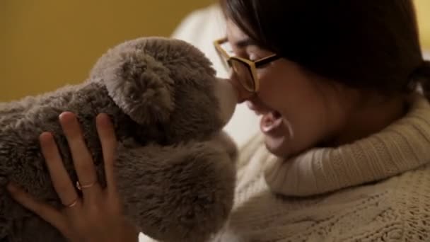 Een jong meisje in een witte trui en dragen bril wordt gespeeld met een zacht stuk speelgoed. Jeugd. Speelgoed — Stockvideo