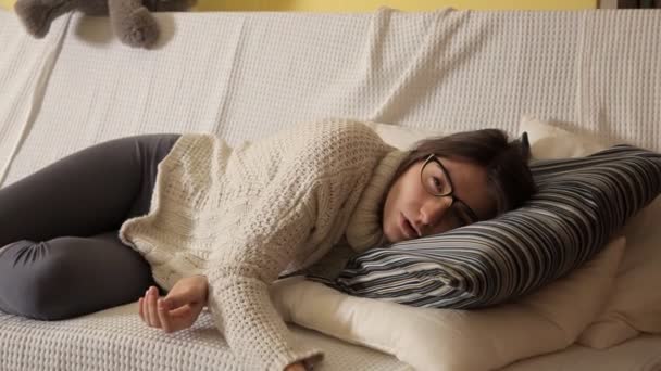 A young girl in a winter evening at home in a white sweater on the couch with glasses watching TV and falling asleep. Evening. House. Comfort. Heat. Winter — Stock Video