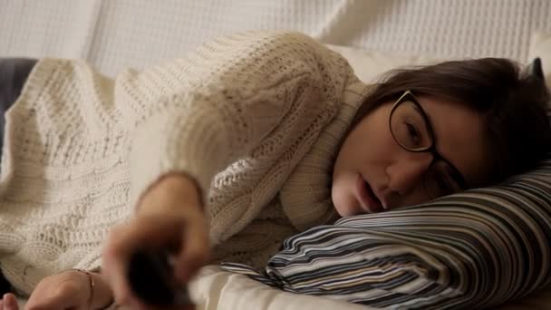 Молода дівчина в зимовий вечір вдома в білому светрі на дивані з окулярами дивиться телевізор і засинає. Вечір. Будинок. Затишок. Тепло. Зима — стокове відео