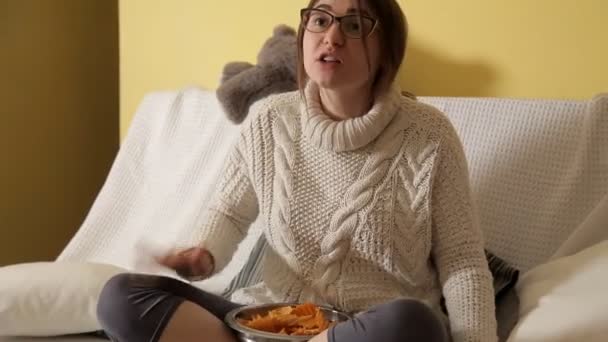 Niña en la noche de invierno en casa en un suéter blanco en el sofá con gafas, viendo la televisión y comiendo patatas fritas. Buenas tardes. Casa. Comodidad. Invierno — Vídeo de stock