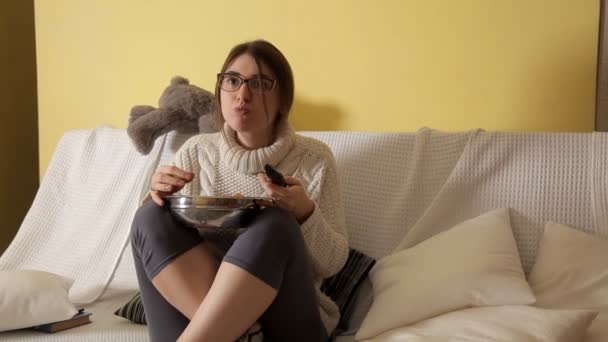 Niña en la noche de invierno en casa en un suéter blanco en el sofá con gafas, viendo la televisión y comiendo patatas fritas. Buenas tardes. Casa. Comodidad. Invierno — Vídeo de stock