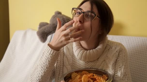 Молодая девушка в зимний вечер дома в белом свитере на диване в очках, смотрит телевизор и ест чипсы. Добрый вечер. Хаус. Комфорт. Зима — стоковое видео