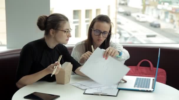 Duas jovens garotas de negócios estão sentadas em um café, comendo macarrão chinês e discutindo o desenvolvimento de negócios. Trabalho. Comer — Vídeo de Stock