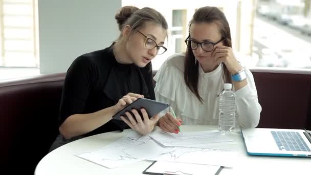 Due ragazze d'affari in occhiali si siedono e discutono affari, usano una targa e un computer portatile, disegnano grafici di sviluppo d'affari, bevono l'acqua. Lavoro. Affari. — Video Stock