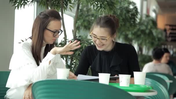 Dos jóvenes empresarias se sientan en una mesa en un café con café y discuten el trabajo. Café, almuerzo, almuerzo, trabajo — Vídeos de Stock