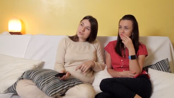 Две молодые девушки сидят дома на диване, в дождливый день, смотрят телевизор и болтают. Дом, дождь, дружба — стоковое видео