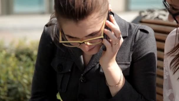 Två unga företag flickor vid lunch sitter i parken nära affärscentret, skrattar och pratar i telefon — Stockvideo