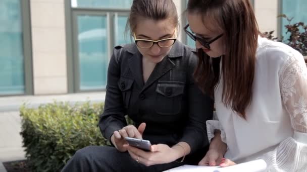 Dos chicas jóvenes de negocios sentadas en el parque cerca del centro de negocios, riendo y mirando las ideas de negocios telefónicas. — Vídeos de Stock