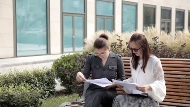 Duas jovens garotas de negócios sentam-se no parque perto do centro de negócios e assistem ao seu plano de desenvolvimento de negócios — Vídeo de Stock