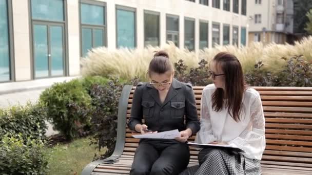 Dos chicas jóvenes de negocios están sentadas en el parque cerca del centro de negocios y esperando una llamada telefónica. — Vídeo de stock