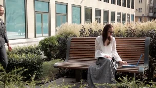 Две молодые деловые девушки летом сидят в парке рядом с бизнес-центром с ноутбуком. Бизнес идеи — стоковое видео