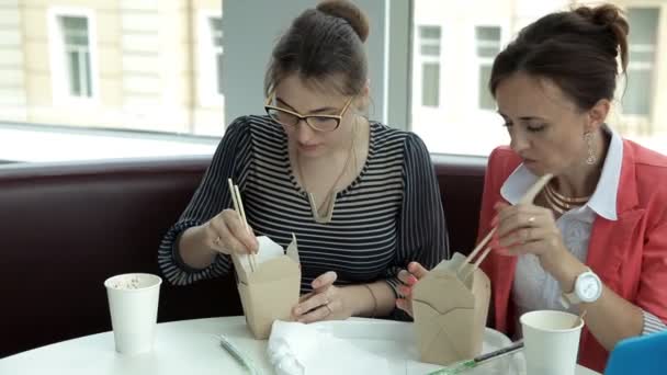 Två unga företag flickor vid lunch sitta och äta läckra nudlar i lådor och dricka kaffe. Lunch på jobbet — Stockvideo