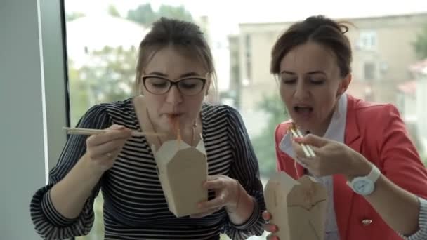 Twee jonge bedrijf meisjes werken bij een Bank en bij het raam Chinese noedels eten lunch en bespreken van bedrijfsontwikkeling — Stockvideo