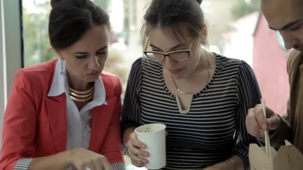 Twee jonge bedrijf meisjes en een jonge zakenman in een business center op lunch noedels eten in vakken en bespreken van bedrijfsontwikkeling — Stockvideo