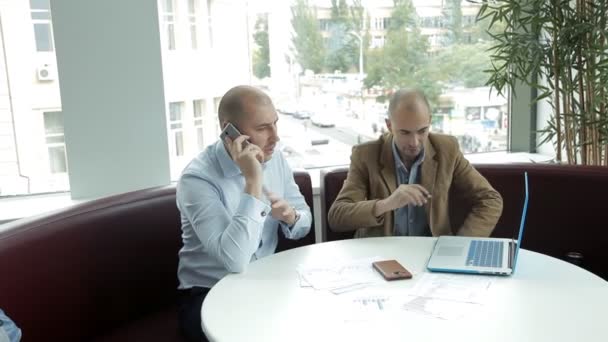 Two businessmen are sitting at a laptop and are talking on the phone by the window — Stock Video