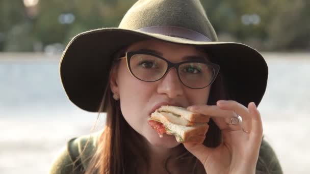 Una joven con gafas y un sombrero está probando un delicioso sándwich. Comida — Vídeo de stock