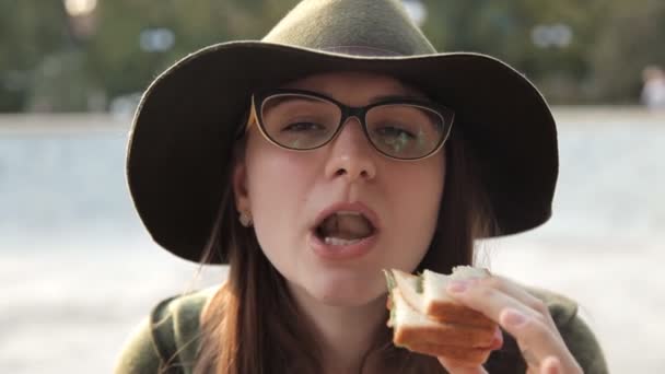 Uma jovem de óculos e um chapéu come uma deliciosa sanduíche. alimentos — Vídeo de Stock