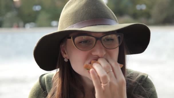 Egy fiatal lány szemüveg és egy kalap egy finom szendvicset eszik. élelmiszer, nem finom, érzelmek — Stock videók