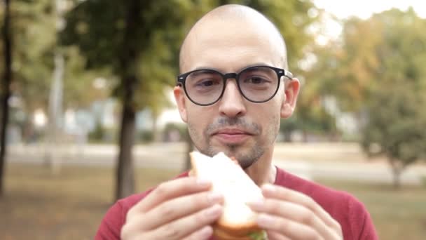 Młody łysy facet w parku zjada kanapkę. Jedzenie nie smaczne, złe sandwich — Wideo stockowe