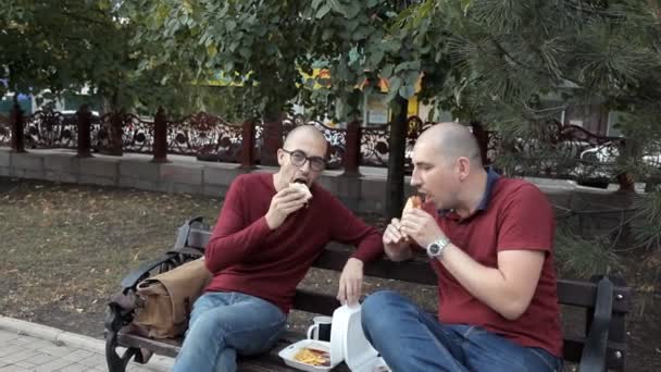 Dos tipos calvos están sentados en el parque comiendo sándwiches y papas fritas. Comida deliciosa, comida rápida — Vídeo de stock