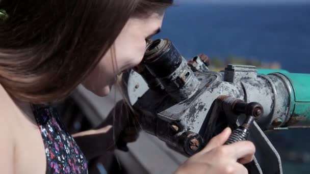 Een jong meisje staat op het observatie dek met uitzicht op de zee en kijkt door een telescoop — Stockvideo