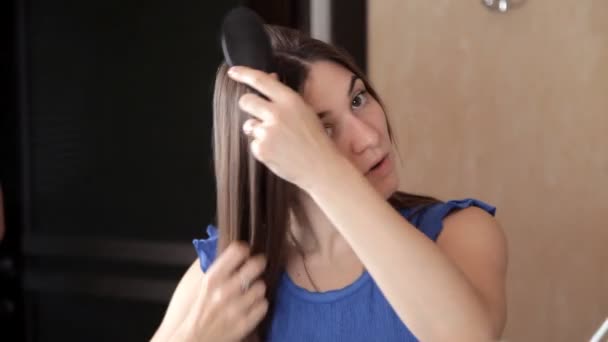 Молодая девушка стоит перед зеркалом и расчесывает длинные волосы. — стоковое видео