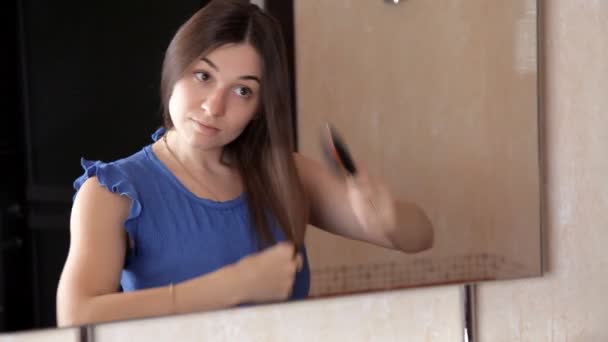 Une jeune fille se tient devant un miroir et peigne ses cheveux longs — Video