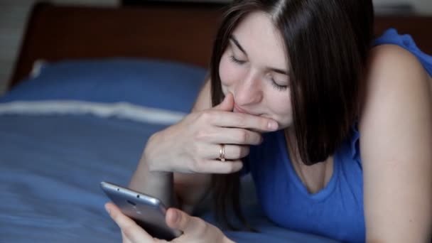 Una joven está acostada en una cama azul con un teléfono y leyendo un mensaje de sms — Vídeos de Stock
