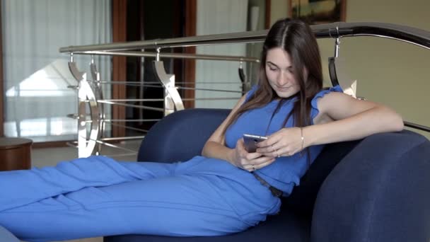 Uma jovem garota se senta no sofá no telefone e lê uma mensagem sms — Vídeo de Stock